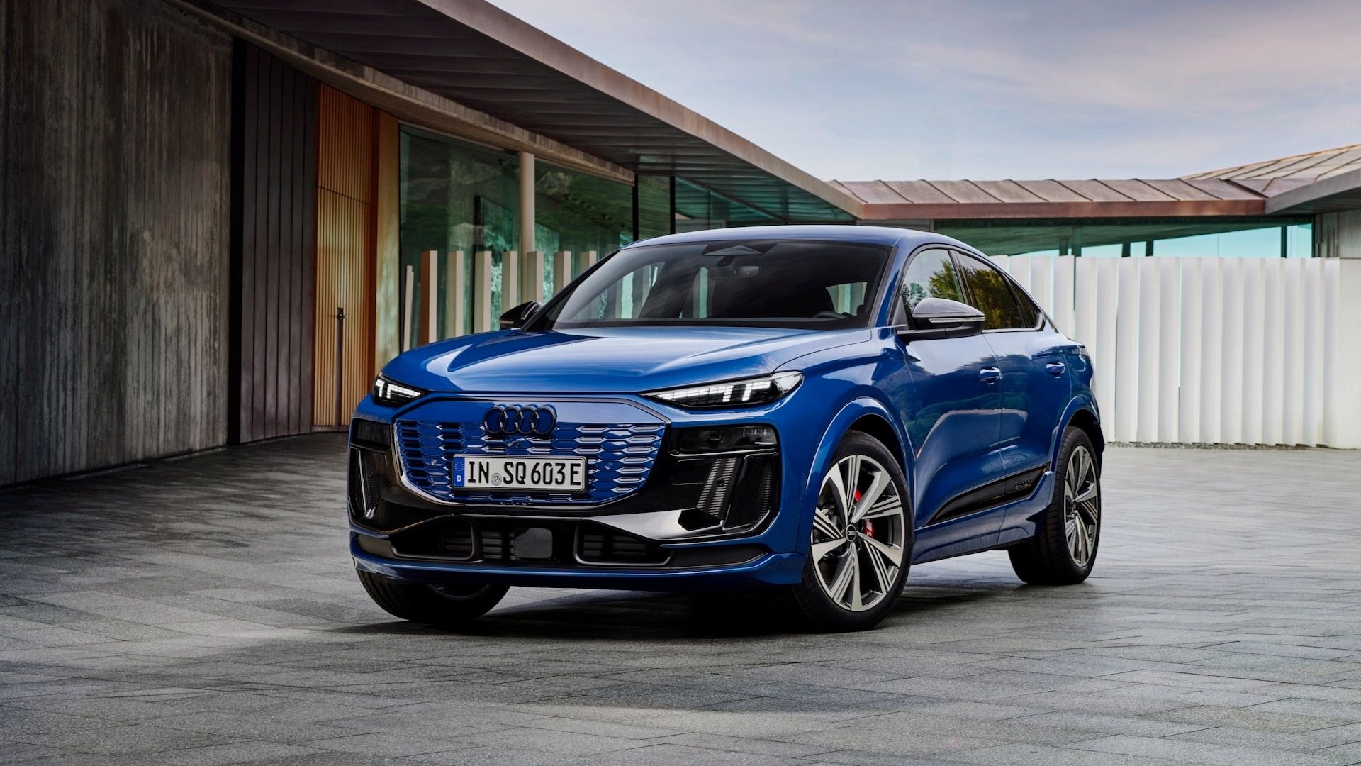 Front three-quarter view of a blue 2024 Audi Q6 Sportback e-tron SUV parked outdoors near modern architecture.