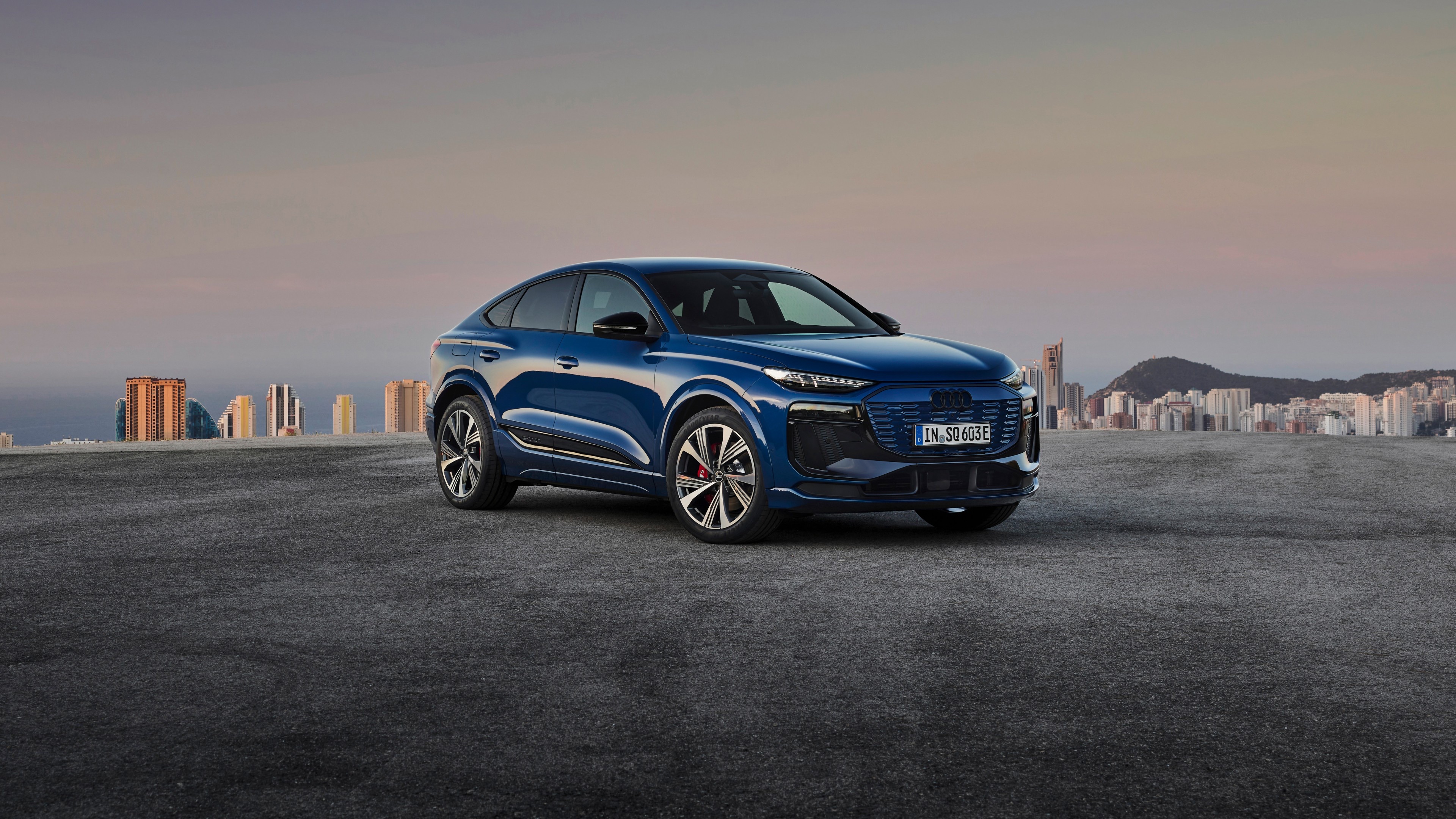 Front three-quarter view of a blue 2024 Audi SQ6 e-tron SUV parked outdoors with a city skyline.