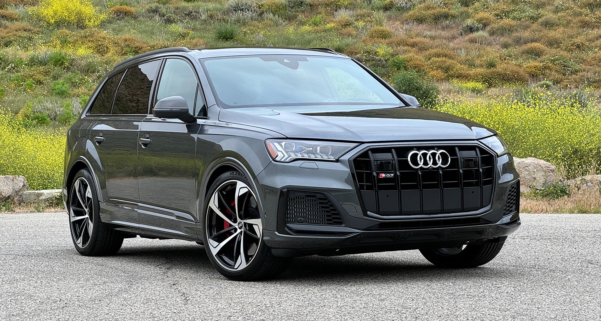 Front three-quarter profile of a grey 2024 Audi SQ7 SUV parked on an asphalt road near a hillside.