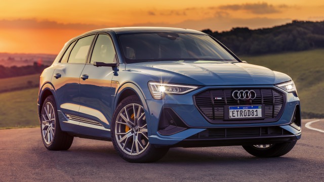 Front three-quarter view of a blue Audi e-tron S SUV parked on a road at sunset.