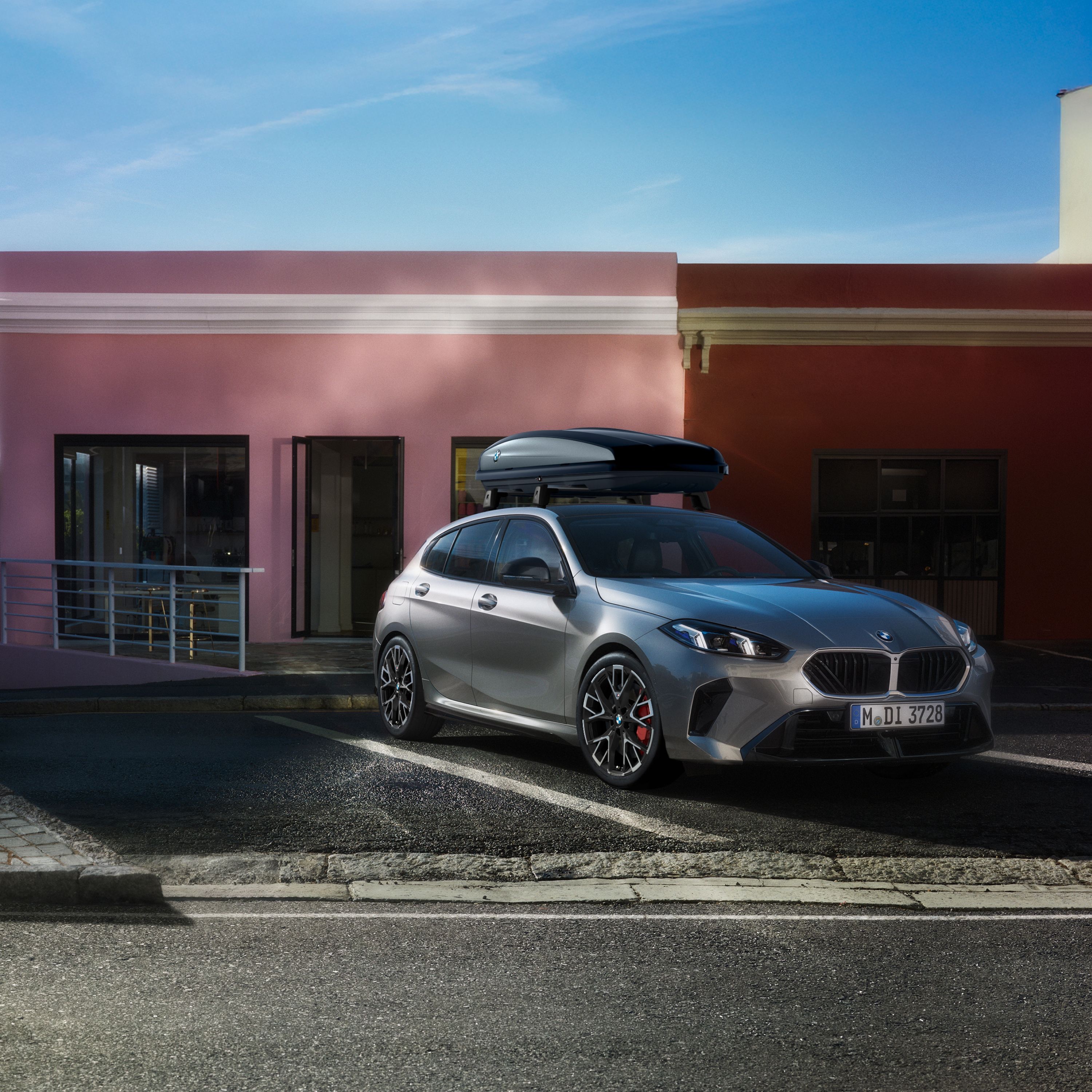 Three-quarter front view of a grey 2024 BMW 1 Series hatchback with a roof box parked outdoors.