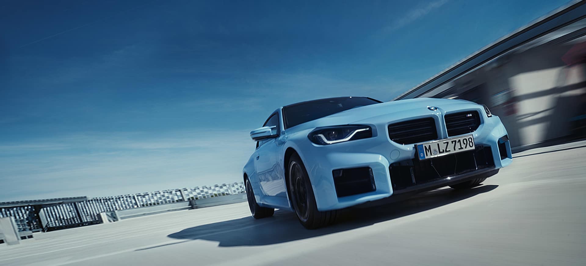 Low-angle front three-quarter view of a light blue 2022 BMW M2 Coupe on a paved rooftop structure.