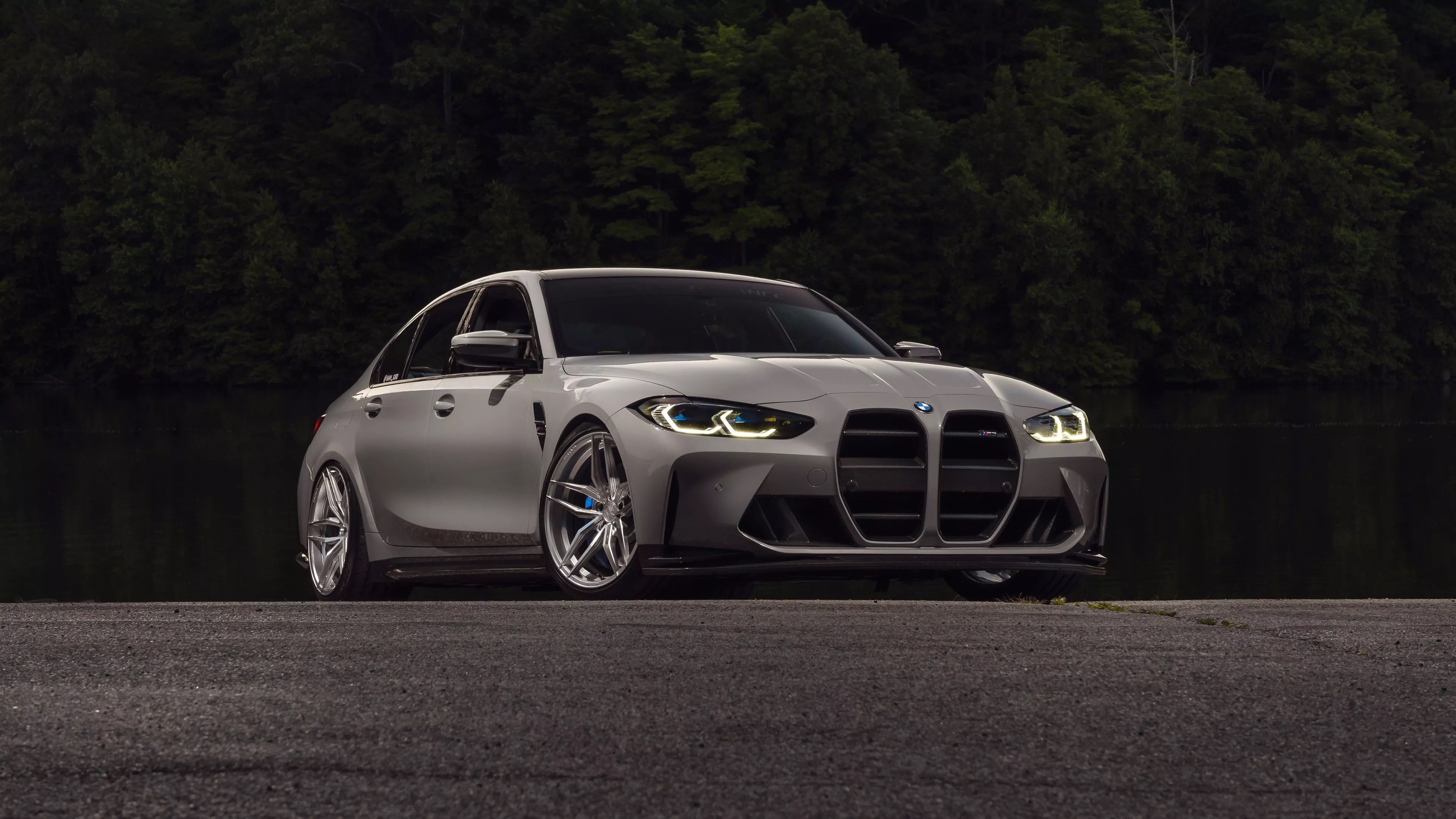 Grey 2020 BMW M3 Sedan shown in a front three-quarter view outdoors near trees and water.