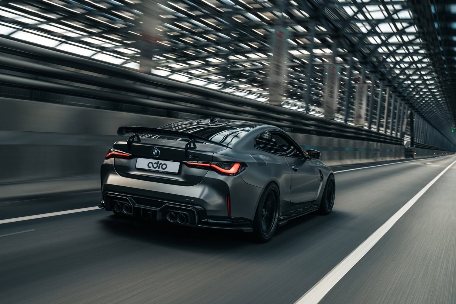 Rear three-quarter view of a dark gray 2020 BMW M4 Coupe driving under a covered structure.