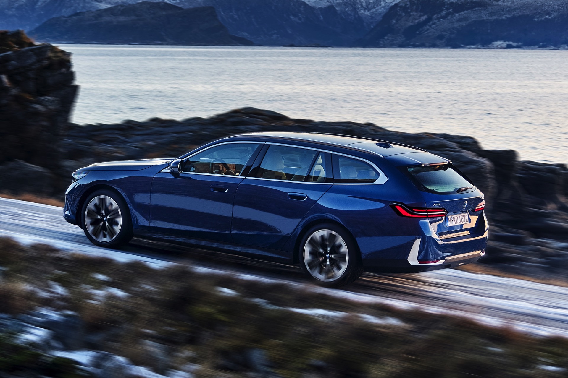 Rear three-quarter view of a blue 2024 BMW 5 Series Touring driving along a coastal road.