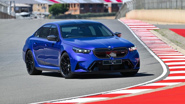 Blue 2024 BMW M5 Sedan driving on a race track with red and white corner kerbing.