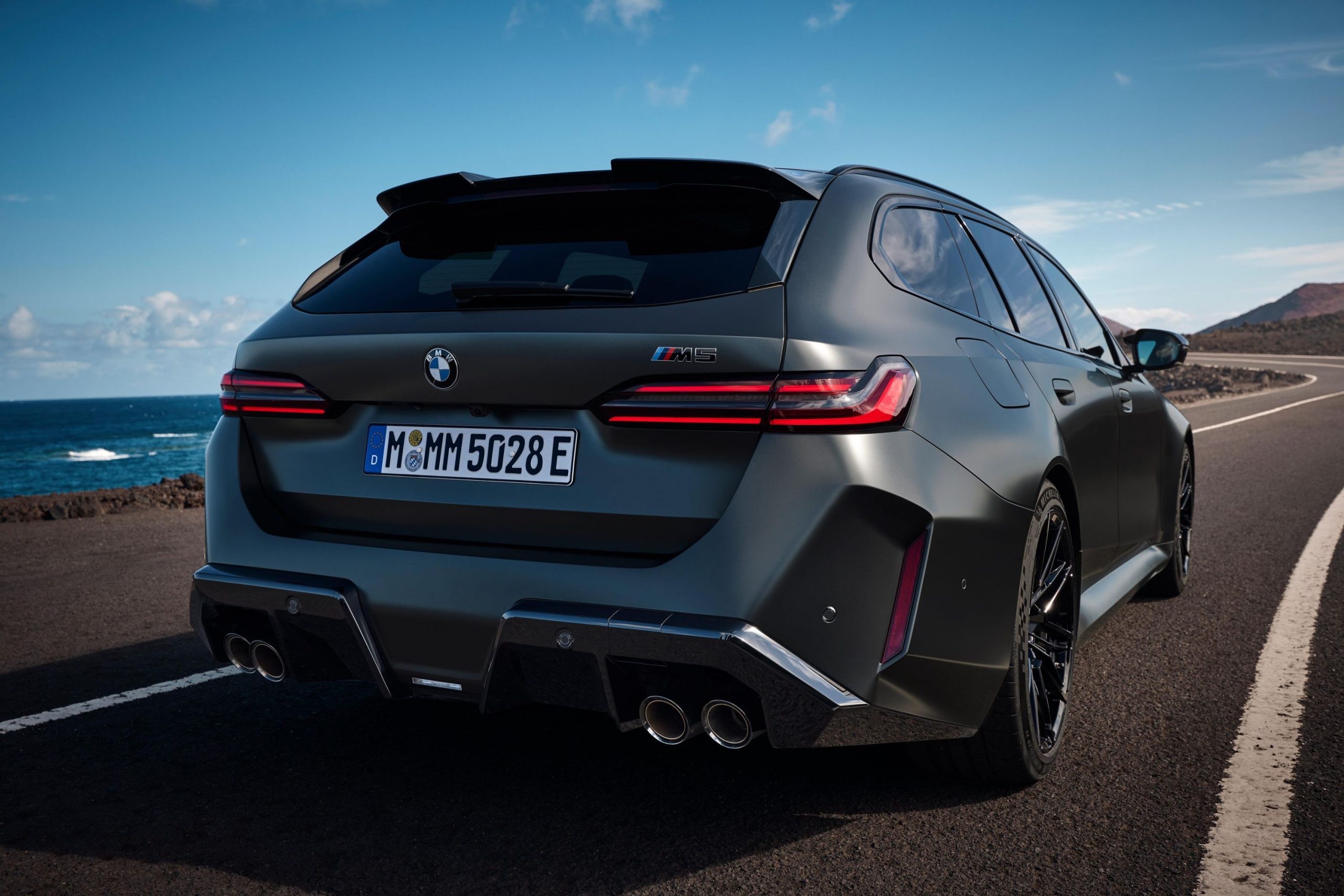 Rear three-quarter of a dark grey 2024 BMW M5 Touring wagon parked along a coastal road.