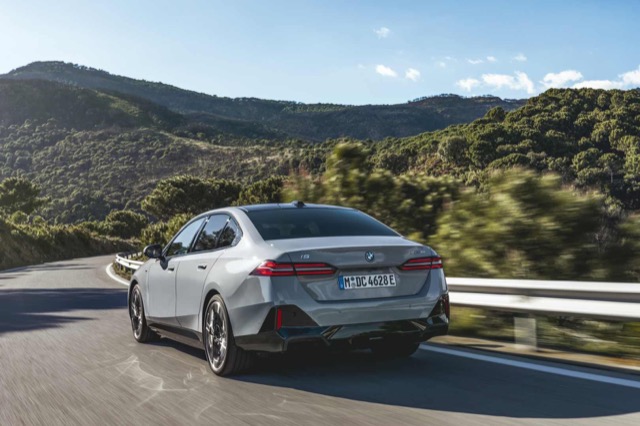 Rear three-quarter view of a grey 2023 BMW i5 sedan driving along a curved mountain road.