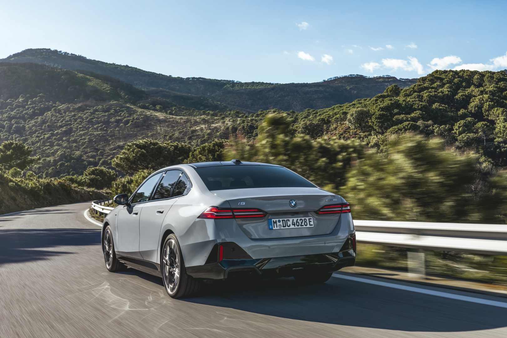 Rear three-quarter view of a grey 2023 BMW i5 sedan driving along a curved mountain road.
