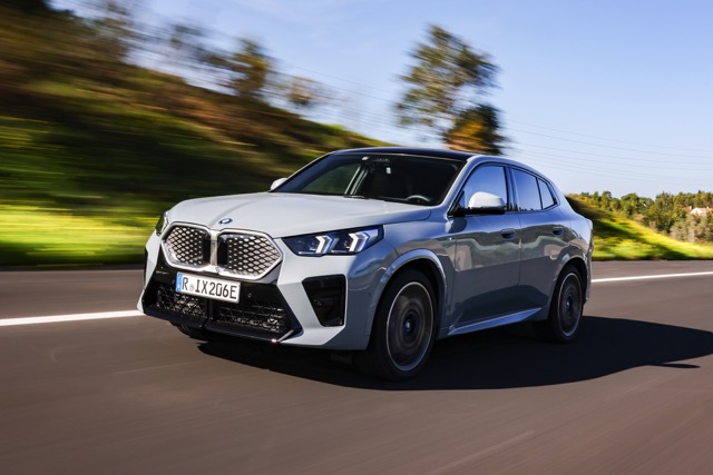 Grey 2023 BMW iX2 SUV driving along a paved road surrounded by trees under a bright blue sky.