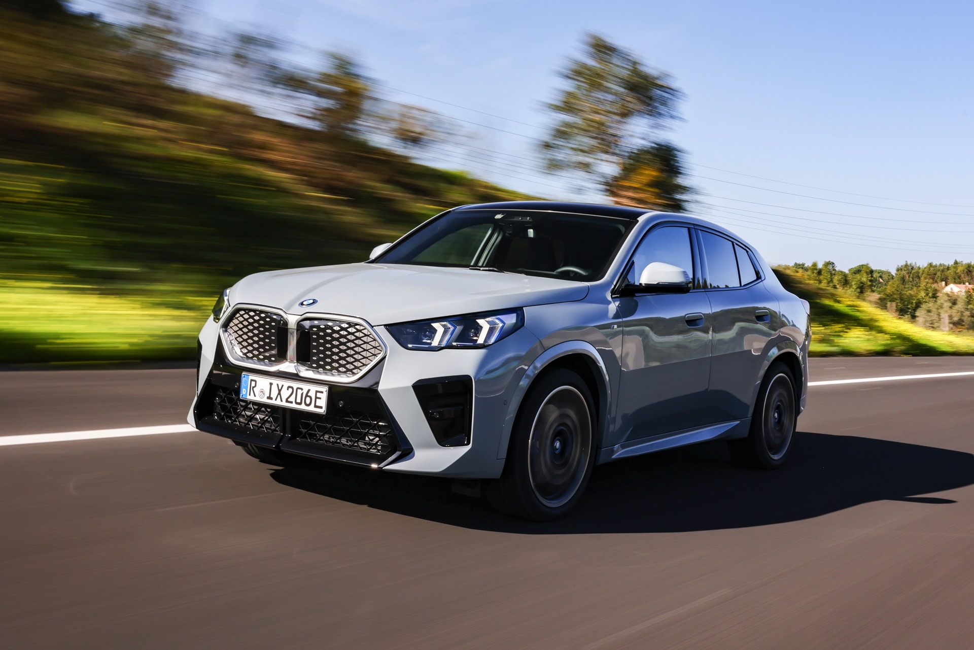 Grey 2023 BMW iX2 SUV driving along a paved road surrounded by trees under a bright blue sky.