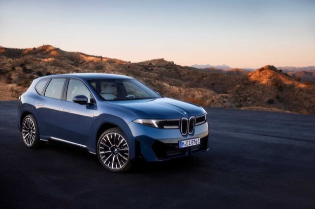 Front three-quarter view of a blue 2025 BMW iX3 SUV parked on an open asphalt road.