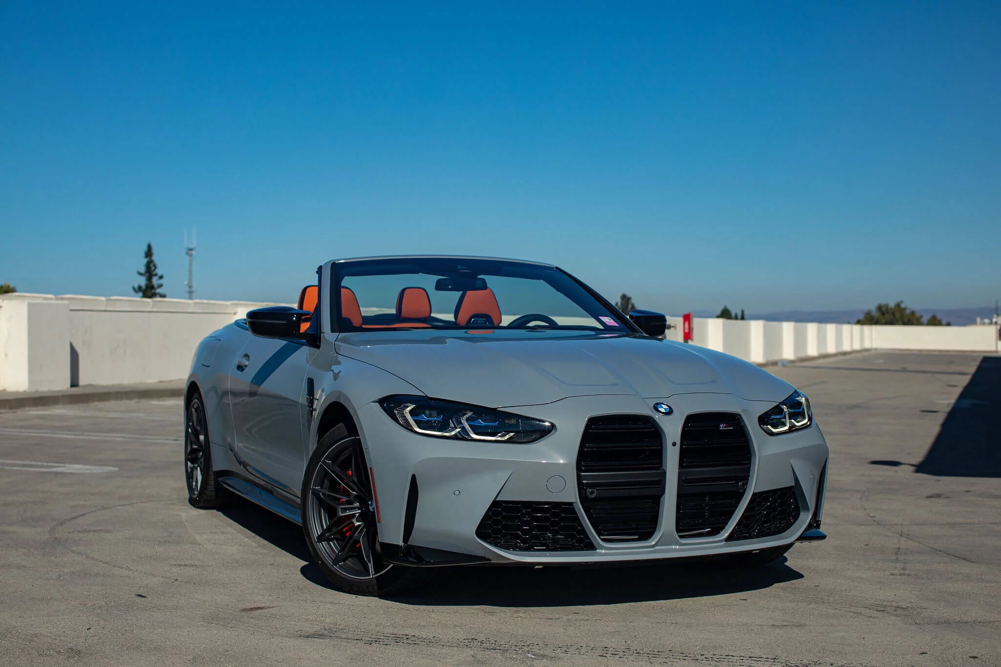 Front three-quarter view of a grey 2021 BMW M4 Convertible with open top on a sunny deck.
