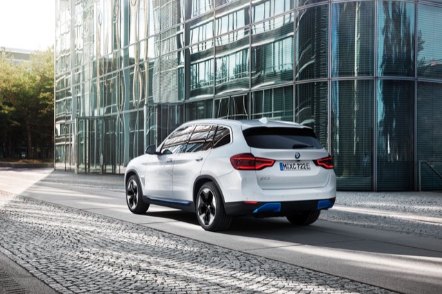 Rear three-quarter view of a white 2020 BMW iX3 SUV parked beside a glass building.