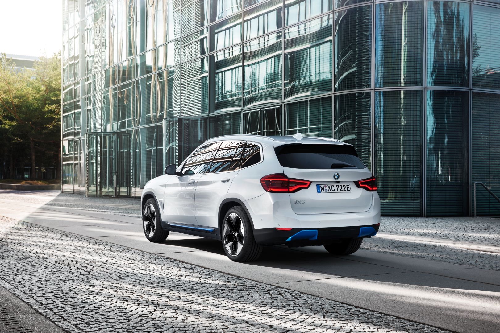 Rear three-quarter view of a white 2020 BMW iX3 SUV parked beside a glass building.