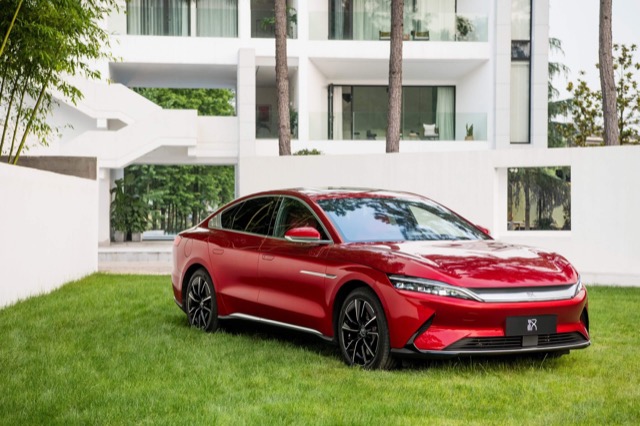 Red 2022 BYD Han EV sedan parked on a grassy lawn outside a white modern building.